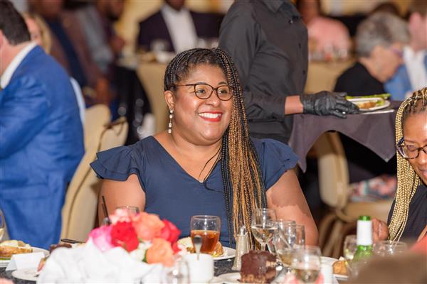 A smiling woman with long braided hair and glasses sits at a decorated dinner table during a formal event. She is wearing a navy blue dress and pearl earrings. The table is set with desserts, drinks, and floral centerpieces. In the background, a server delivers a plate of food and other guests are s